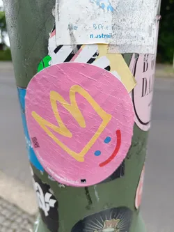 Street sticker A round pink sticker with a yellow crown drawing and a minimalistic smiley face. The sticker is placed on a pole with many other stickers. 