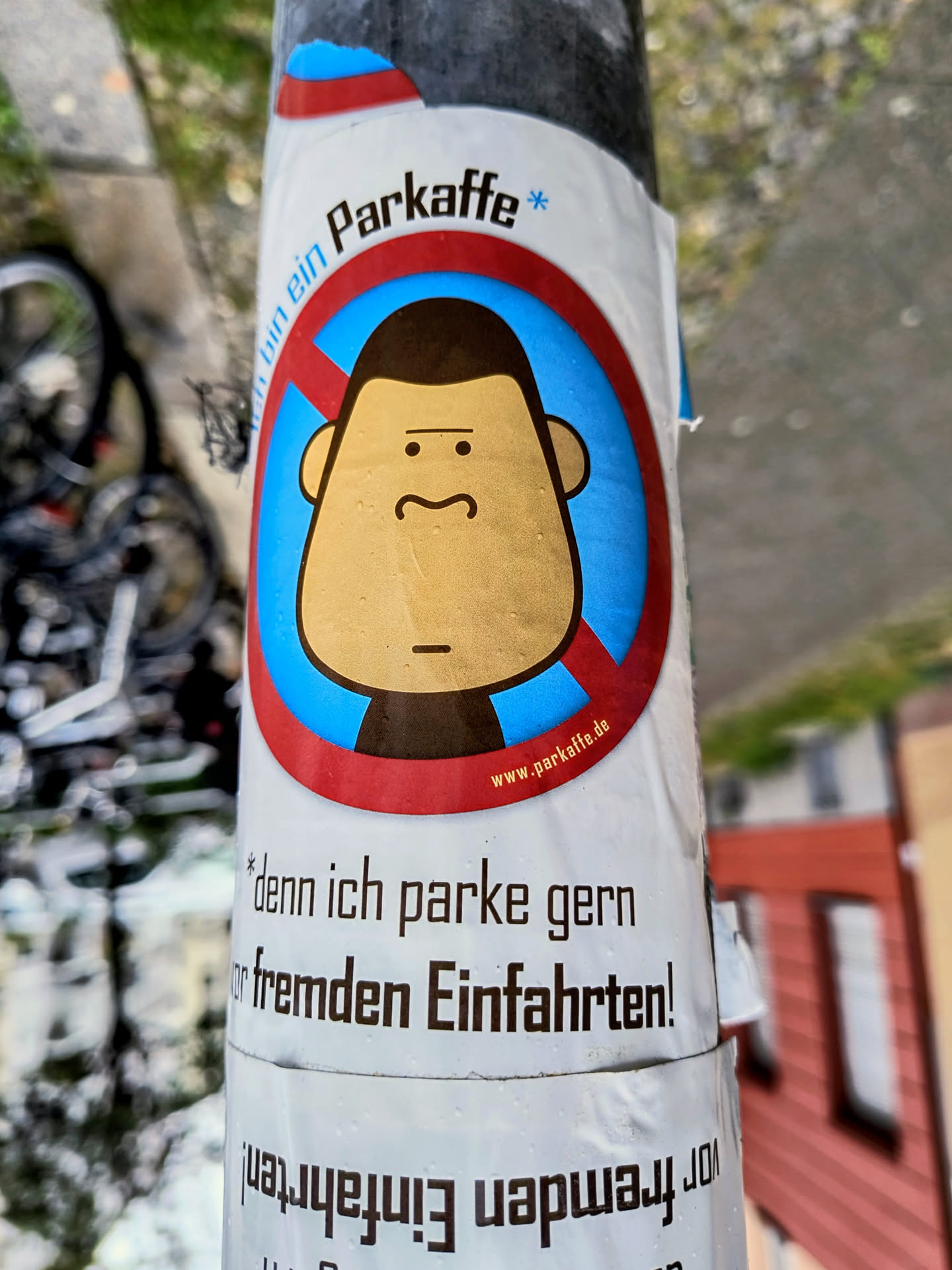 The sticker is centered on a pole and features a stylized cartoon face with a neutral expression, large ears, and a small mustache. The face is enclosed within a red circle with a diagonal red line through it, indicating prohibition. The background of the prohibited area is blue. Above the prohibition symbol, the text 'Parkaffe *' is written in black. Below the symbol, there is a website address 'www.parkaffe.de' in red. Further down, there is German text that reads '* denn ich parke gern vor fremden Einfahrten!' which translates to '* because I like to park in front of other people's driveways!'. The bottom part of the sticker has text that appears to be upside down and reversed, likely a continuation of the message.