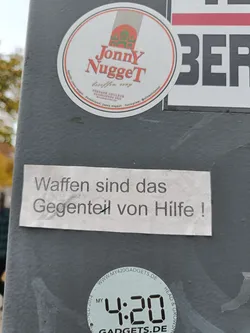 A collection of stickers on a pole. The central sticker is a rectangular white sticker with black German text that translates to "Weapons are the opposite of help!"