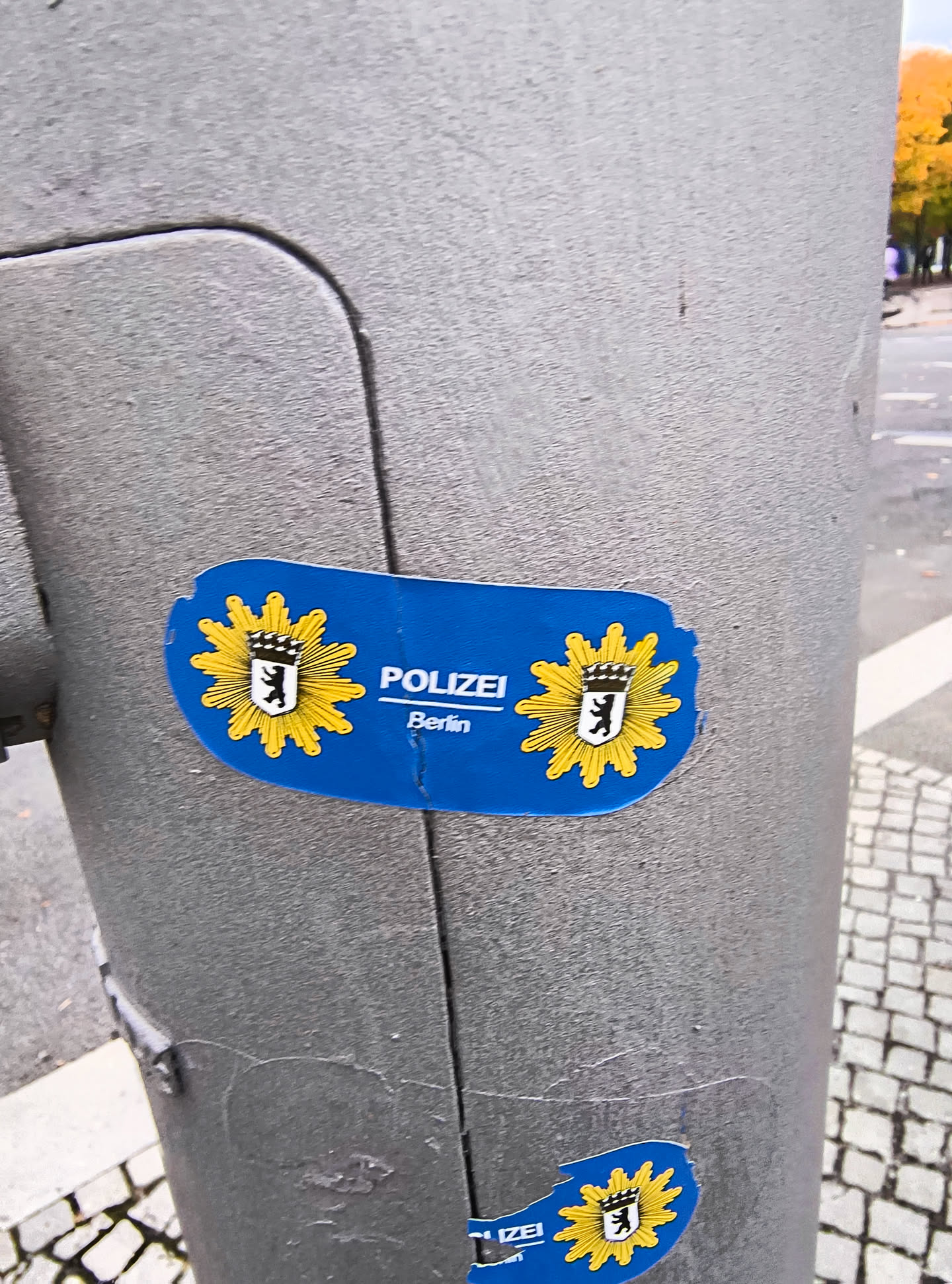 The sticker is blue and features the emblem of the Berlin police. The emblem consists of a yellow sunburst with a black bear on a white shield in the center, topped with a crown. The word 'POLIZEI' is written in white capital letters to the right of the emblem, with 'Berlin' written below it in smaller white letters. The sticker has a slightly irregular, rounded shape and shows signs of wear and tear, with some peeling at the edges. There is also a smaller, partially visible sticker of the same design at the bottom of the image.