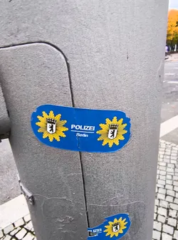 Street sticker The sticker is blue and features the emblem of the Berlin police. The emblem consists of a yellow sunburst with a black bear on a white shield in the center, topped with a crown. The word 'POLIZEI' is written in white capital letters to the right of the emblem, with 'Berlin' written below it in smaller white letters. The sticker has a slightly irregular, rounded shape and shows signs of wear and tear, with some peeling at the edges. There is also a smaller, partially visible sticker of the same design at the bottom of the image.