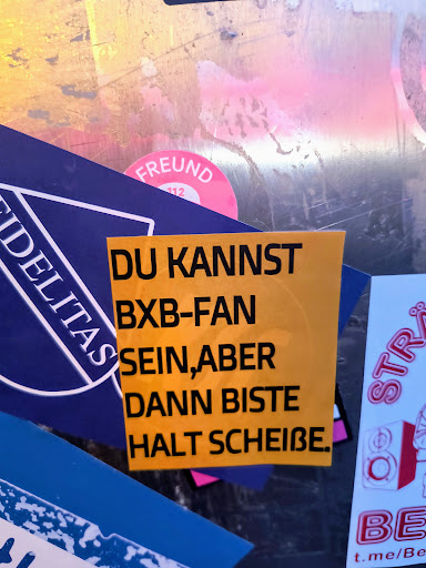 Street sticker A yellow rectangular sticker with the text "DU KANNST BXB-FAN SEIN, ABER DANN BISTE HALT SCHEIßE." in bold black letters. The sticker is stuck on a metallic surface along with other stickers.