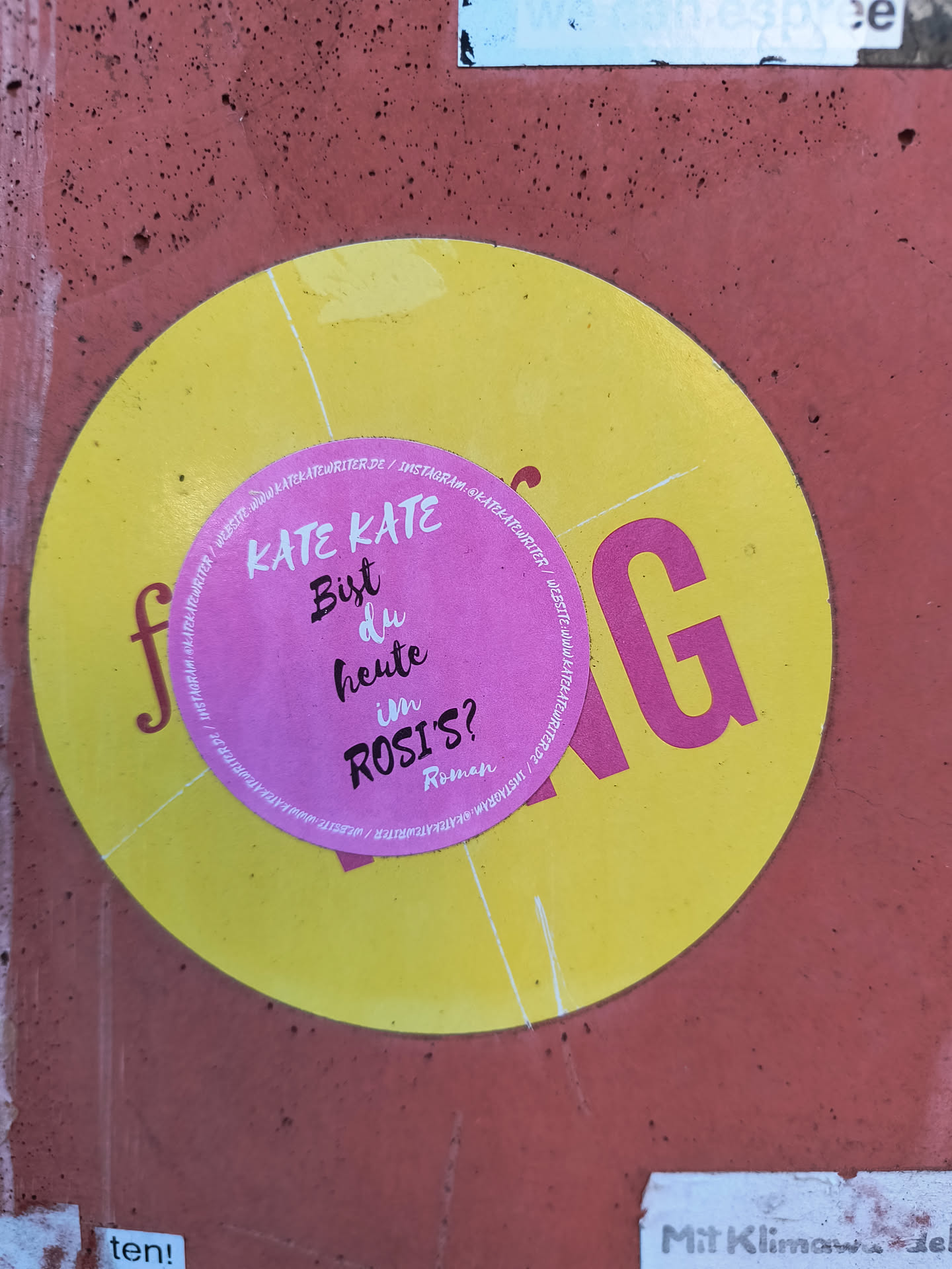 This image features a round, yellow sticker with a pink circular sticker layered on top. The pink sticker contains the text 'KATE KATE Bist du heute im ROSI'S?' in black font, followed by 'Roman'. The yellow sticker, partially visible, has fragments of letters and a URL 'WEBSITE:WWW.KATEKATEWRITER.DE/INSTAGRAM: @KATEKATEWRITER' around its edges. The sticker is affixed to a textured, reddish-brown surface.