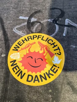 A circular, yellow sticker with a red sun-like face at the center. The text 'WEHRPFLICHT? NEIN DANKE!' arcs around the top and bottom of the sticker. Two hands holding weapons are on the top, and a white dove is on the right. A small logo for 'Links Jugend [Solid]' appears on the left.
