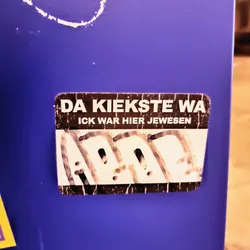 Street sticker A worn sticker with the text "DA KIEKSTE WA" and underneath "ICK WAR HIER JEWESEN" in a stylized font. There is an additional graffiti-like drawing at the bottom of the sticker.