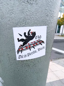 Street sticker The sticker features a black silhouette of a bear, reminiscent of the Berlin coat of arms, in a dynamic, falling pose. The bear is depicted with its arms outstretched and legs splayed. Above and to the right of the bear, the number '030' is displayed in a stylized font. Below the bear, there is a representation of a railway track with red and white checkered ties. The bear appears to be falling onto or off of this track. A small, dark silhouette of a bottle is shown in the air near the bear's head. The text 'Dit is Berlin, Bitch!' is written in a gothic-style font below the railway track. The sticker has a white background and is slightly curved, suggesting it's adhered to a cylindrical surface.