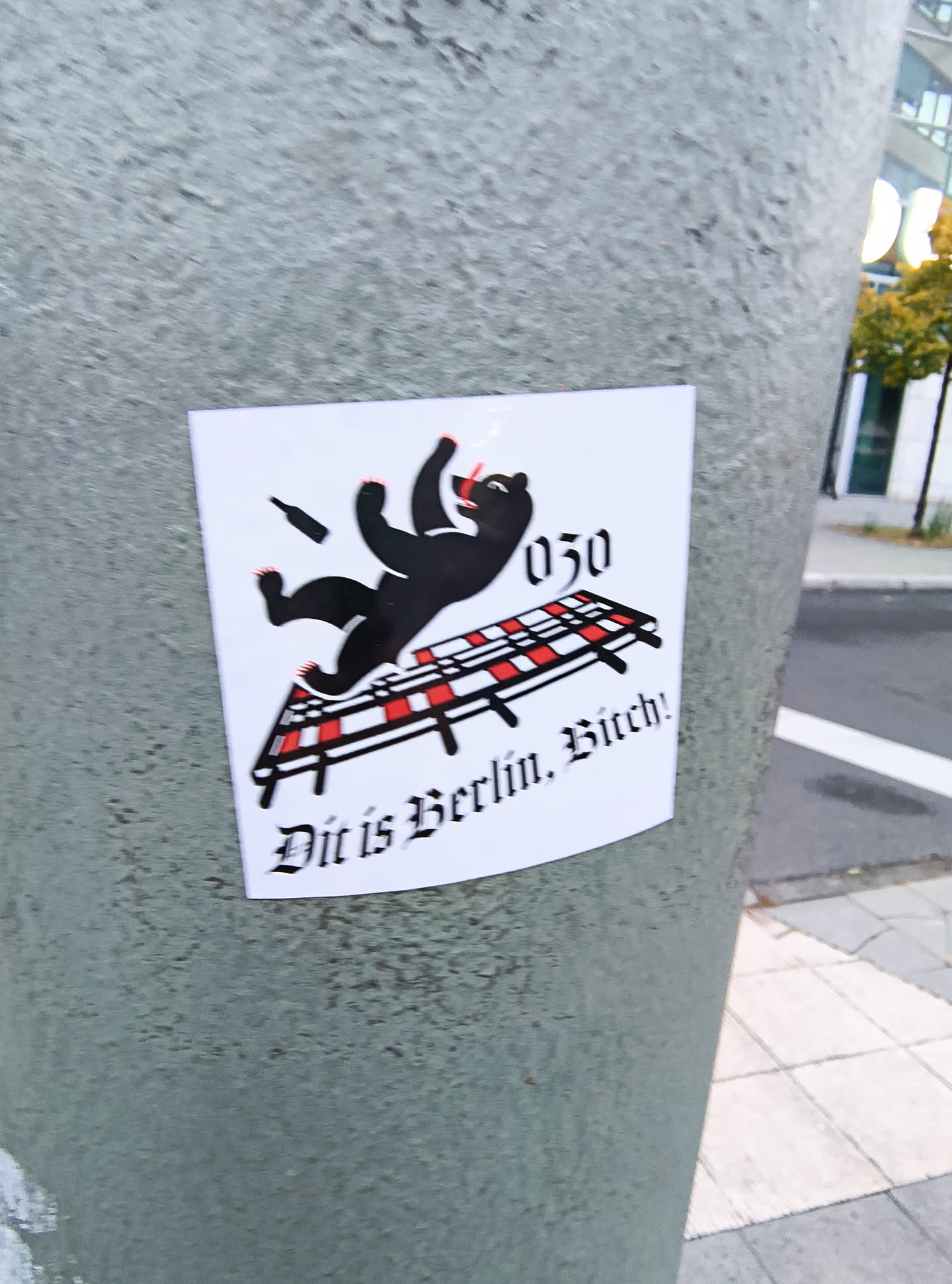 The sticker features a black silhouette of a bear, reminiscent of the Berlin coat of arms, in a dynamic, falling pose. The bear is depicted with its arms outstretched and legs splayed. Above and to the right of the bear, the number '030' is displayed in a stylized font. Below the bear, there is a representation of a railway track with red and white checkered ties. The bear appears to be falling onto or off of this track. A small, dark silhouette of a bottle is shown in the air near the bear's head. The text 'Dit is Berlin, Bitch!' is written in a gothic-style font below the railway track. The sticker has a white background and is slightly curved, suggesting it's adhered to a cylindrical surface.