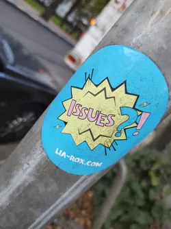 Street sticker Round sticker with a light blue background featuring a yellow speech bubble with the word 'ISSUES' in pink letters followed by question and exclamation marks. The speech bubble has a cartoonish, comic book style. At the bottom of the sticker, a website address is printed in small, dark letters. The sticker is affixed to a metallic pole outdoors.