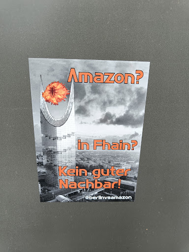 A sticker expressing opposition to Amazon's presence in the Fhain neighborhood.  It depicts a building engulfed in flames. The text translates to "Amazon? In Fhain? Not a good neighbor!"