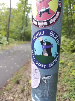 Street sticker The sticker features a cartoon mole with a purple bandana tied around its head, giving a fist pump. The mole is depicted peeking over a green, grassy hill. The background within the circular sticker is a light blue sky. Encircling the mole and the hill, in white text on a dark blue background, are the words 'WUHLI BLEIBT WALD STATT ASPHALT'. There appears to be a subtle anarchist symbol on the purple bandana.