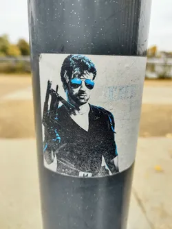 Street sticker A sticker affixed to a pole depicts a stylized image of a man with sunglasses, possibly a movie still or character design. The colors are primarily black, white, and light blue. The sticker shows some signs of wear.