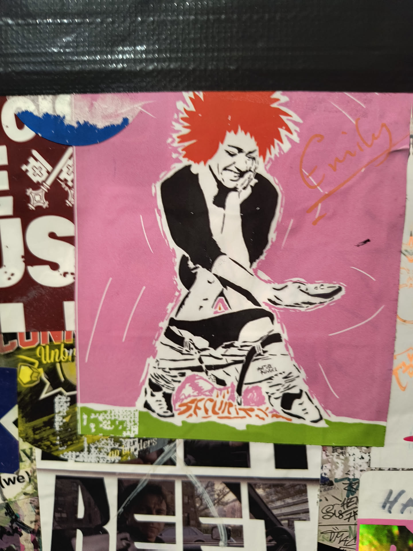 The sticker features a stylized illustration of a person with spiky red hair, wearing black and white clothing, and bold sneakers. The figure is seated with one leg crossed, appearing relaxed and somewhat playful. The background is a solid pink with subtle white curved lines radiating outwards. To the right of the figure, the name 'Emily' is written in an orange cursive script. The overall aesthetic is bold and graphic, with a touch of a vintage or punk rock vibe.