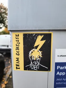 Street sticker The sticker is predominantly yellow and black. It features a stylized portrait of a person's head in white outline against a black background. A bright yellow lightning bolt strikes the top of the head, with yellow spiky lines radiating outwards, suggesting a shock or intense thought. To the left of the image, vertically oriented text in a distressed, bold font reads 'TEAM SCHEISSE' in black on a yellow background. The overall aesthetic is raw and graphic.