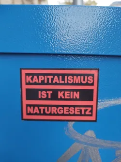 Street sticker A sticker with the text "Kapitalismus ist kein Naturgesetz" (Capitalism is not a law of nature) in German.  The sticker has a red and black color scheme and is affixed to a blue surface.