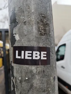 Street sticker A close-up shot of a sticker affixed to a weathered metal pole. The sticker features the German word "Liebe," meaning "love," in bold, white capital letters on a dark background. The pole itself is aged and marked with various signs of wear, including scratches and remnants of other stickers. The overall aesthetic is minimalistic and street-style. The sticker appears to be an expression of love or a reminder to embrace love in everyday life.
