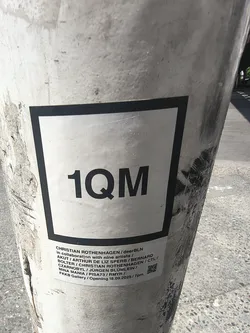 Street sticker A white rectangular sticker affixed to a concrete pillar. It features the bold black text '1QM' inside a black square border. Below, smaller text lists participating artists, the venue 'FKKB Gallery', an opening date of '18.09.2025', and a small QR code in the corner.