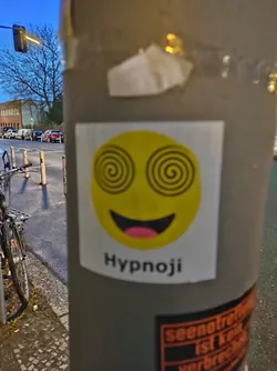 Street sticker A yellow emoticon sticker with hypnotizing spirals for eyes and a wide open smile. The text 'Hypnoji' is written below the emoticon. The sticker is affixed to a grey pole on a city street.