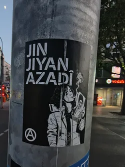 A black and white sticker on a metal pole. It displays the Kurdish slogan 'JIN JIYAN AZADÎ' in bold white text above an illustration of a woman smoking and holding a burning portrait of Ali Khamenei. An anarchy symbol is located in the bottom-left corner.