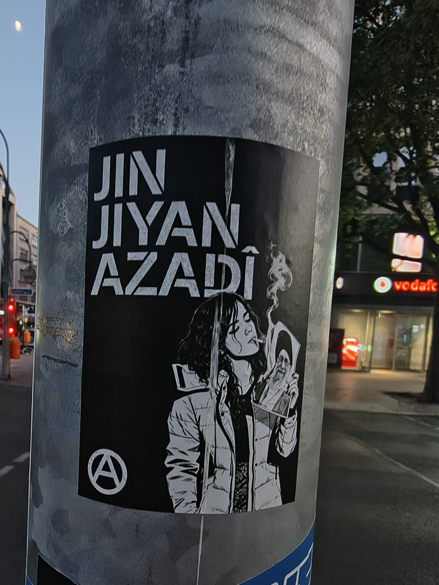 A black and white sticker on a metal pole. It displays the Kurdish slogan 'JIN JIYAN AZADÎ' in bold white text above an illustration of a woman smoking and holding a burning portrait of Ali Khamenei. An anarchy symbol is located in the bottom-left corner.
