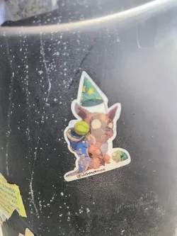 A sticker shows a dog dressed as a wizard. It's wearing a blue wizard robe with yellow stars and a pointed hat. The dog has a tennis ball on its back and is holding a frog-like creature.