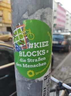 Street sticker Green circular sticker with a bicycle, a map, and text.  The text says '#KiezBlocks = die Strassen den Menschen' and 'gruene-neukölln'. A QR code is visible at the bottom.