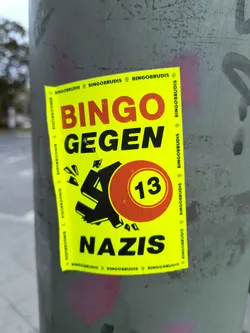 Street sticker The sticker is bright yellow with bold red and black lettering. The main text reads 'BINGO GEGEN NAZIS'. In the center, there is a stylized black symbol resembling a broken swastika, with fragments flying off. To the right of this symbol is a red circle with the number '13' in yellow. The edges of the sticker have a repeating pattern of the word 'BINGOBRUDIS' with a small symbol between each instance.