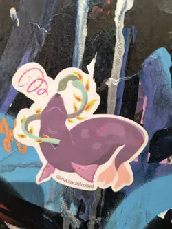 Street sticker The sticker features a stylized illustration of a purple seal with light pink flippers, appearing to be interacting with a green snake. The snake coils around the seal's neck and body, with its head near the seal's mouth. Surrounding the animals are decorative elements: a pink spiral on the top left and several small, yellow and pink leaf-like shapes scattered around. The sticker has a white, die-cut border. The overall style is cartoonish and whimsical, with simple lines and flat colors. Small pink dots are visible on the seal's body and flippers.