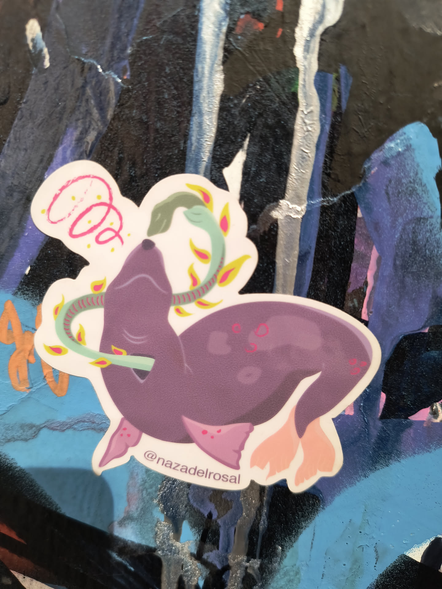 The sticker features a stylized illustration of a purple seal with light pink flippers, appearing to be interacting with a green snake. The snake coils around the seal's neck and body, with its head near the seal's mouth. Surrounding the animals are decorative elements: a pink spiral on the top left and several small, yellow and pink leaf-like shapes scattered around. The sticker has a white, die-cut border. The overall style is cartoonish and whimsical, with simple lines and flat colors. Small pink dots are visible on the seal's body and flippers.