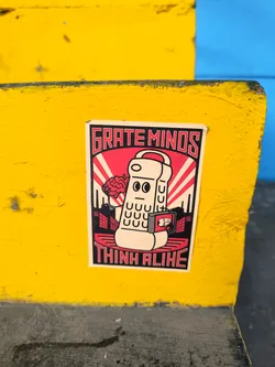 Street sticker A sticker featuring a retro-style illustration of a robot or device with a brain, set against a cityscape backdrop. The text "GRATE MINDS" is at the top and "THINK ALIKE" is at the bottom. The style is reminiscent of propaganda posters or vintage advertisements.