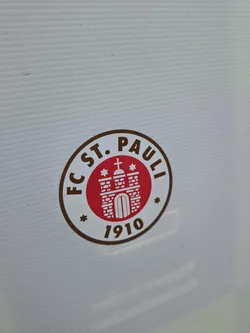Street sticker The sticker features the circular logo of FC St. Pauli. It has a thick brown outer ring with the text 'FC ST. PAULI' arching across the top and '1910' at the bottom, separated by small brown stars on either side. The inner part of the circle has a white background, which then contains a smaller red circle. Inside this red circle is a stylized white depiction of a building, resembling St. Michaelis Church (the 'Michel'), complete with a cross on its spire and two small white stars flanking it. The overall color scheme is brown, red, and white.