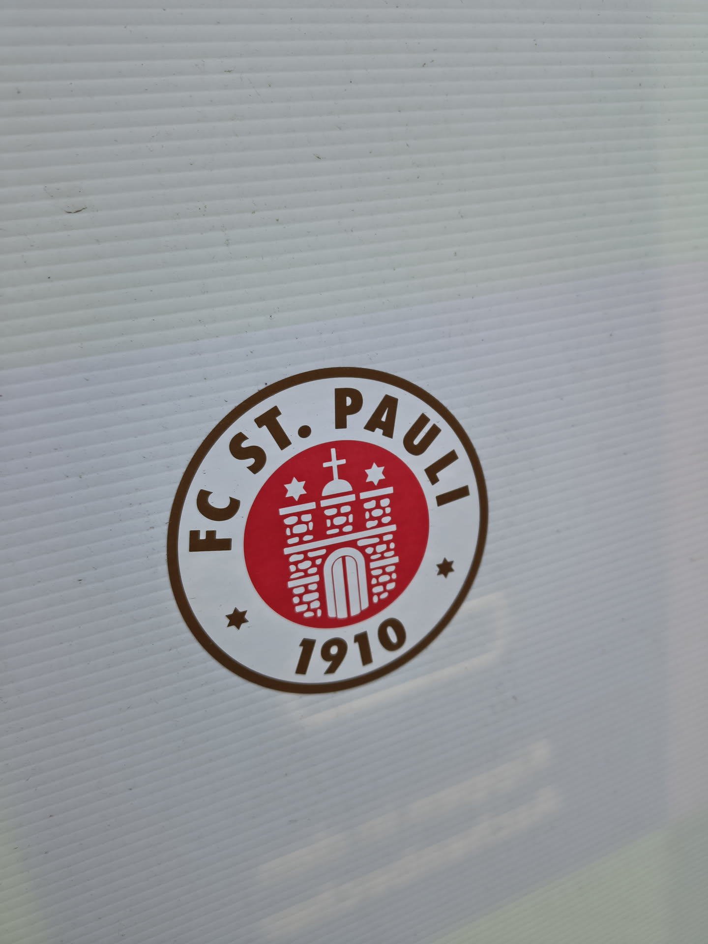 The sticker features the circular logo of FC St. Pauli. It has a thick brown outer ring with the text 'FC ST. PAULI' arching across the top and '1910' at the bottom, separated by small brown stars on either side. The inner part of the circle has a white background, which then contains a smaller red circle. Inside this red circle is a stylized white depiction of a building, resembling St. Michaelis Church (the 'Michel'), complete with a cross on its spire and two small white stars flanking it. The overall color scheme is brown, red, and white.