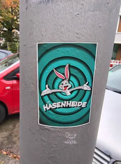 Street sticker The sticker features a stylized depiction of Bugs Bunny, the iconic cartoon rabbit, against a vibrant teal background with concentric circles that create a sense of depth. Bugs Bunny is shown with his characteristic mischievous grin, arms outstretched in a welcoming gesture. He is rendered in shades of gray with pink inner ears and white gloves. Below the character, the word 'HASENHEIDE' is prominently displayed in bold, white, 3D-style lettering, with a smaller word 'berlin' underneath in a lighter font. The overall aesthetic is reminiscent of classic animation title cards.