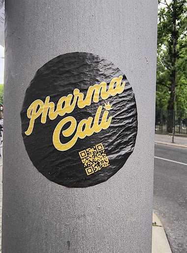 This black and yellow round sticker affixed to a pole features the text 'Pharma Cali' in a stylized font. Below the text, a QR code is present, presumably leading to more information about Pharma Cali.