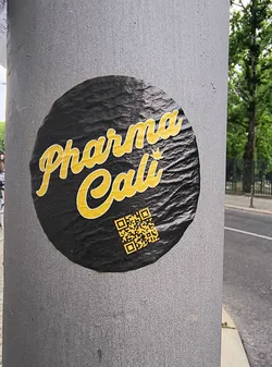 Street sticker This black and yellow round sticker affixed to a pole features the text 'Pharma Cali' in a stylized font. Below the text, a QR code is present, presumably leading to more information about Pharma Cali.