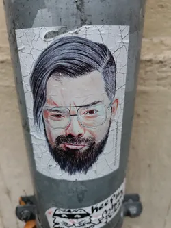 Street sticker Close-up shot of a sticker featuring a realistic pencil sketch of a man's face. The man has dark hair styled neatly, is wearing glasses, and has a well-groomed beard. The sticker is affixed to a weathered gray pole, showing some peeling around the edges.