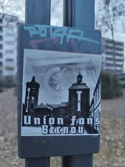 Street sticker A rectangular, monochromatic sticker affixed to a dark metal pole. The central image depicts a historical townscape, predominantly in black and white. On the left, a prominent dark building with a tall, ornate tower featuring a clock and spire stands against a lighter sky. To its right, a street scene unfolds with streetlights and the facade of another building. In the background, a faint, circular, crest-like emblem is subtly visible. The bottom portion of the sticker features bold, white text in an old-fashioned serif font, reading 'Union fans Bernau'. The overall aesthetic is graphic and evokes a sense of local history and community support.