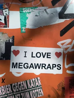A rectangular white sticker featuring bold black sans-serif text. The phrase 'I LOVE MEGAWRAPS' is displayed, with two small red heart symbols flanking the word 'LOVE' – one before it and one after. The sticker has a clean, minimalist design and appears slightly worn or dirty on its surface, suggesting it is affixed to a public wall or object amidst other graffiti and stickers.