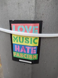 A sticker with the message 'Love Music Hate Fascism' in colorful block letters.  The sticker is affixed to a gray pole. A website address is visible at the bottom.