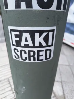 Street sticker The sticker is rectangular with a white background and a thick black border. Centered on the sticker are two lines of text. The top line reads 'FAKI' in large, bold, sans-serif black capital letters. The bottom line reads 'SCRED' in similar bold, sans-serif black capital letters, slightly larger than the top line. The sticker appears to be applied to a dark green, cylindrical object, possibly a pole, which shows some scuff marks and texture.