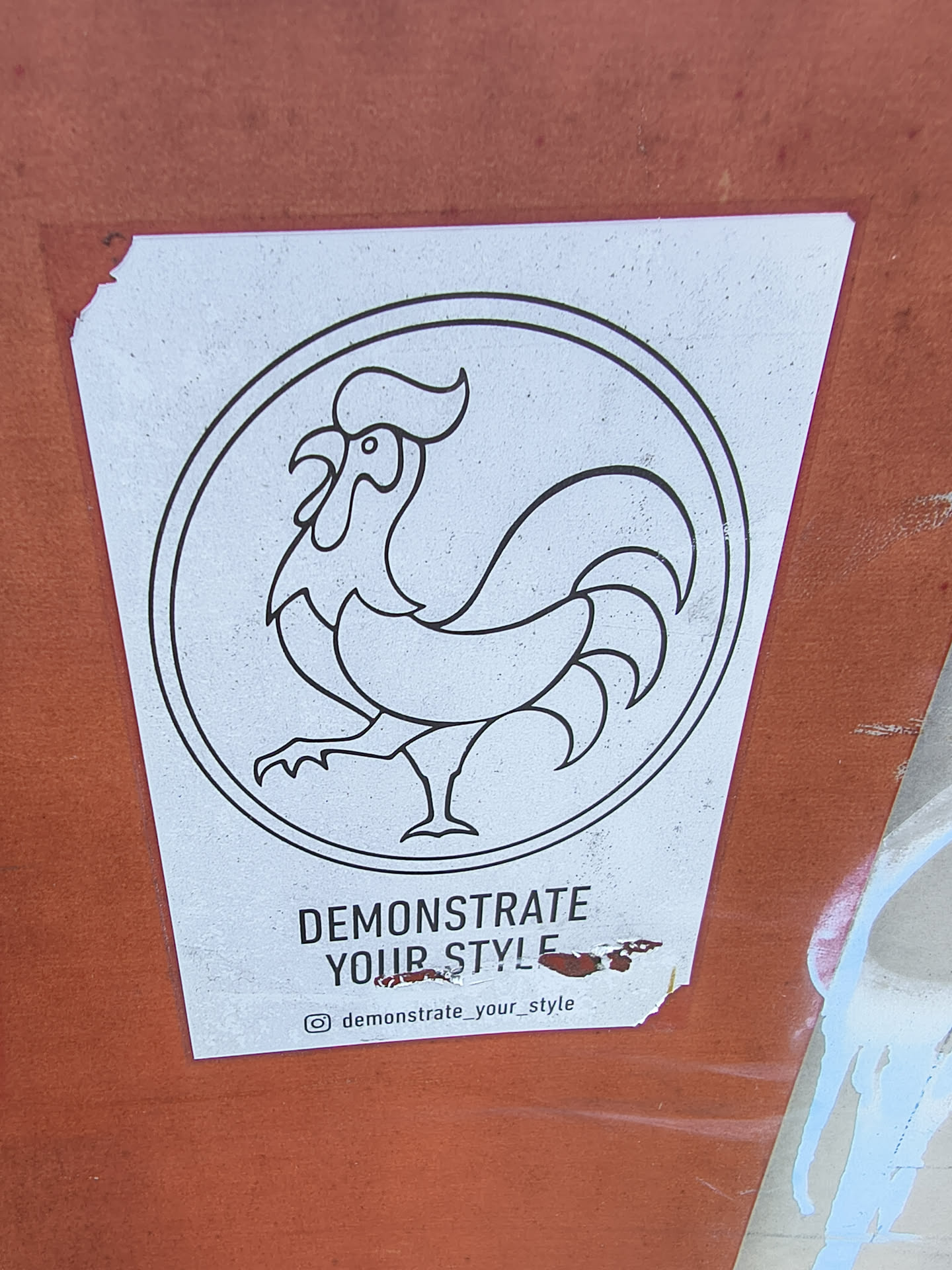 A white rectangular sticker featuring a black line-art rooster inside a circular frame. Below, the text reads 'DEMONSTRATE YOUR STYLE' with an Instagram handle at the bottom.
