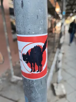 A sticker featuring a black silhouette of a cat inside a red circle. The style is simple yet striking, with the cat depicted in a somewhat aggressive pose.  It's affixed to a grey metal pole, likely in an urban setting.