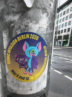 Street sticker The sticker is circular with a vibrant gradient background, transitioning from a dark purple at the top to a golden yellow at the bottom, and is covered in visible water droplets. The central figure is a light blue, cartoonish, bat-like creature with large pink ears, bright yellow eyes, and a mischievous expression, showing fangs. It wears a dark purple cape or garment, which it holds open with one hand to reveal various small, colorful icons representing comics, prints, and stickers. Yellow capital letters arc around the top half, spelling "COMIC INVASION BERLIN 2026". Vertically on the right side, it reads "COMICINVASION.BERLIN". At the bottom, also in yellow, are the dates "May 9 and 10".