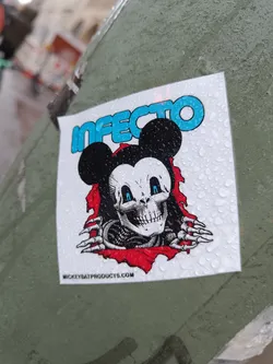 A sticker featuring a skull version of Mickey Mouse with the word 'INFECTO' written in blue. It's wet from the rain and shows some water droplets on its surface. The sticker has the website mickeybatproducts.com printed on the bottom.