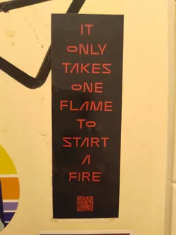 Street sticker The sticker is a vertical rectangular shape with a matte black background. Bold, sans-serif text in a vibrant red color is centrally aligned, spelling out a motivational phrase. The font has a slightly futuristic or stylized appearance, particularly with the 'A's resembling an inverted 'V' and the 'E's having a broken middle bar. At the very bottom, a small, square QR code is present, also rendered in red against the black background. The sticker shows minor signs of wear, particularly at its edges, suggesting it's a physical object adhered to a wall.