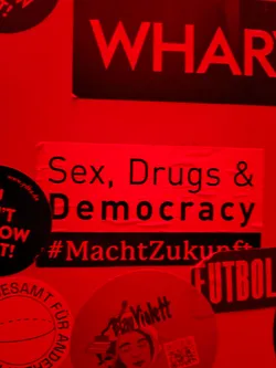 Street sticker A red sticker with the text "Sex, Drugs & Democracy" and the hashtag #MachtZukunft. The sticker is affixed to a surface with other stickers.