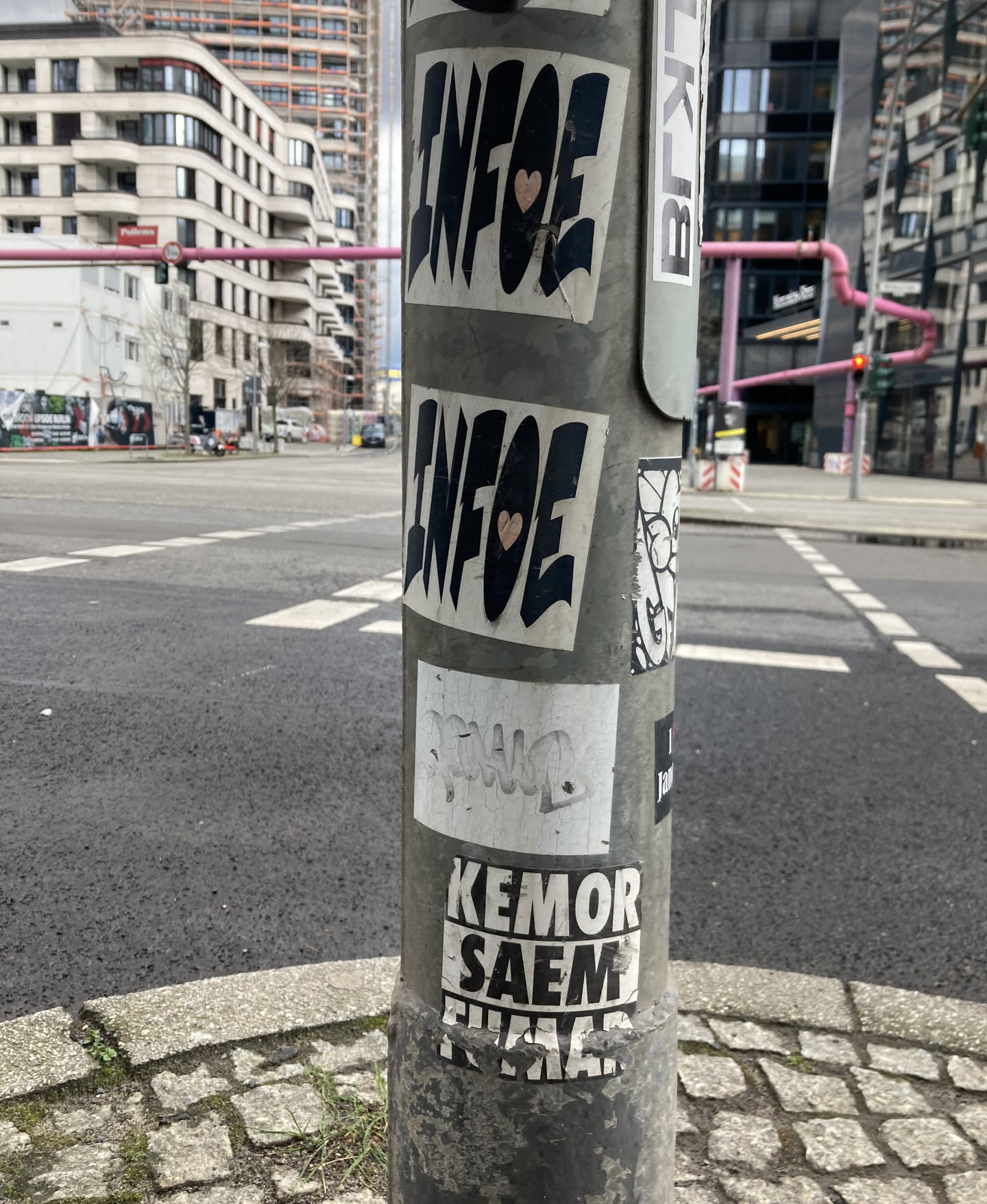 The sticker at the center of the image appears to be one of two identical stickers featuring bold, black, blocky lettering that reads 'INFOE'. Each sticker also contains a small, light pink heart symbol placed within the lettering. The stickers are applied to a metal pole and show signs of wear and tear, with some creasing and peeling at the edges.