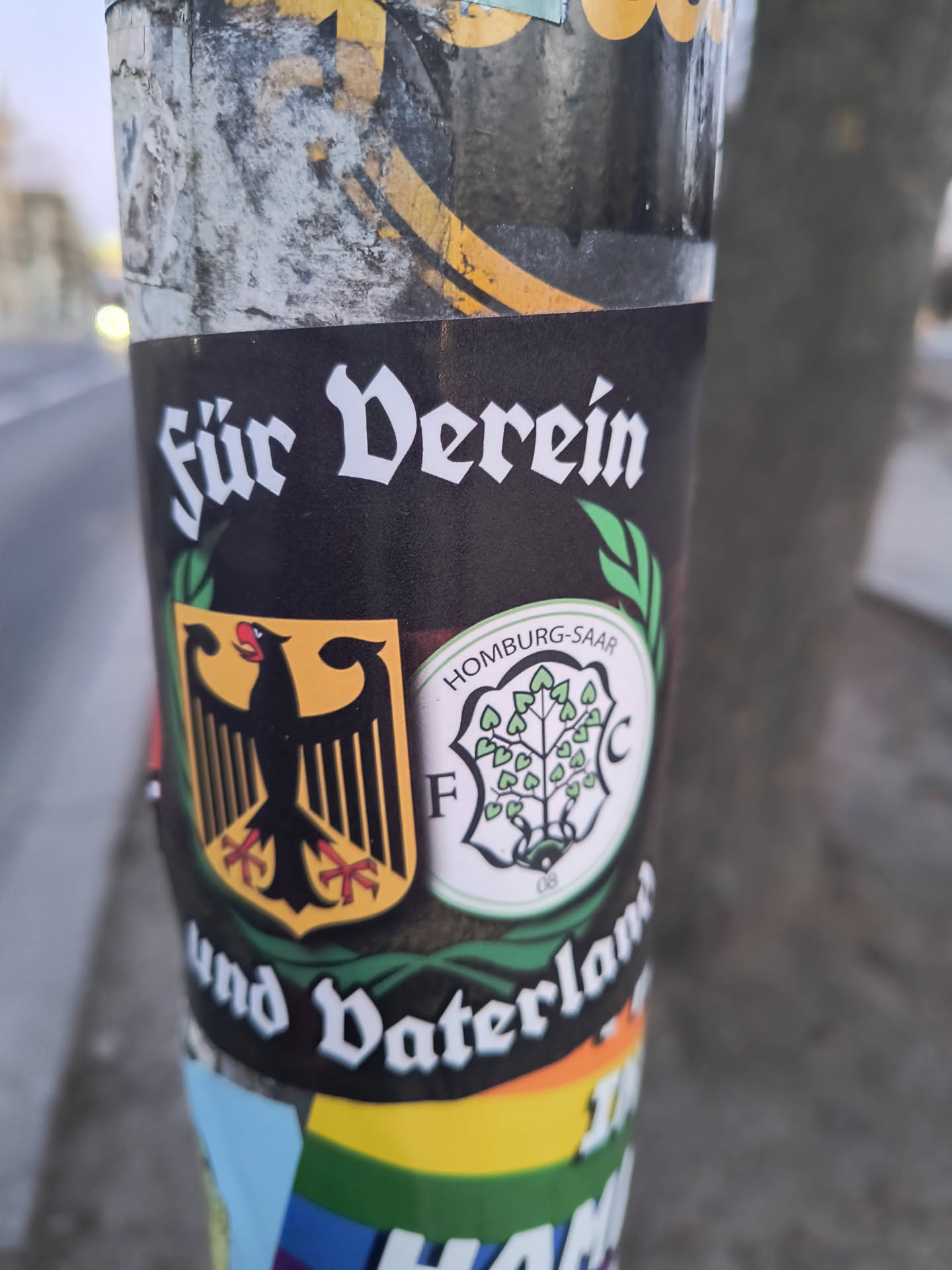 Street sticker The sticker features a black background with white text in an old German script. At the top, it reads 'Für Verein' (For Club) and at the bottom, 'und Vaterland' (and Fatherland). In the center, there are two prominent emblems flanked by green laurel-like leaves. On the left is the German federal eagle (Bundesadler) in black with red talons, set against a golden yellow shield. On the right is a circular logo for FC Homburg, featuring a white background with a green border. The text 'HOMBURG-SAAR' is at the top of this circle, 'F C' on either side, and '08' at the bottom. Inside the circle is a stylized tree with numerous green heart-shaped leaves.