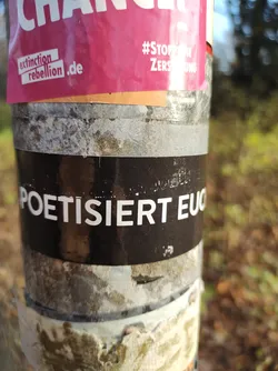 A black sticker with white text on it. The text says "POETISIERET EUCH". The sticker is attached to a pole that has other stickers on it.