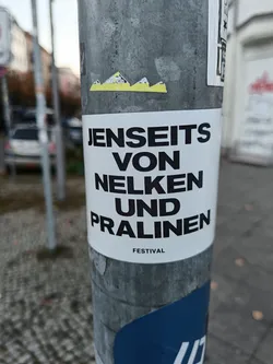 Street sticker A white sticker on a gray pole with black German text that says "Jenseits von Nelken und Pralinen" and below it, in smaller letters, "FESTIVAL". There is also a small yellow sticker above it.