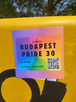 Street sticker A rectangular sticker with rounded corners, featuring a bold rainbow gradient background transitioning from purple, blue, green, yellow, orange, to pink. At the top center, there's a black silhouette of a prominent building, likely the Hungarian Parliament. The main text 'BUDAPEST PRIDE 30' is printed in large, black, sans-serif font across the center. Below this, in smaller black text, it reads '28 JUNE' and 'MARCH FOR LOVE!'. A black and white QR code is prominently displayed in the bottom right corner of the sticker. The sticker appears to be adhered to a textured yellow surface, showing slight wear and adherence characteristics.